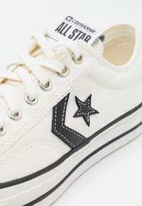 Baskets Converse All Star blanches avec logo étoile noire et chevron, œillets métalliques, lacets blancs et semelle en caoutchouc à rayures noires.