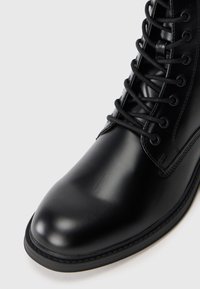 Bota de cuero negro con un acabado suave, punta redonda y cordones negros resistentes. La suela está detallada con costuras en contraste.