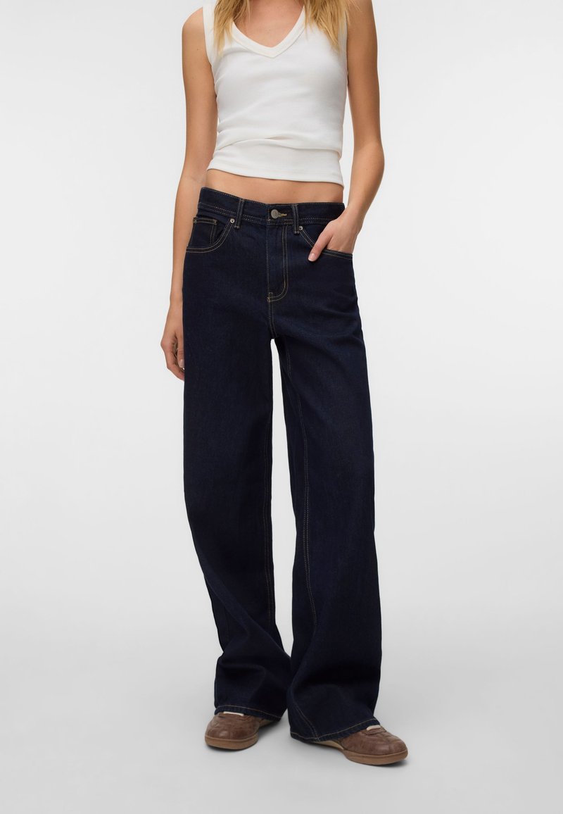 Femme portant un jean large foncé, un top blanc sans manches et des chaussures marron, debout avec une main dans la poche sur un fond uni.