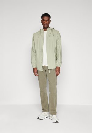 Chaqueta con capucha verde claro y botones a presión, llevada sobre una camiseta blanca. Combinada con pantalones de pana beige y zapatillas blancas.