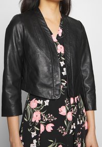 Černá kožená zkrácená bunda s tříčtvrtečními rukávy, s hladkou texturou a minimalistickým designem, nošená přes šaty s květinovým vzorem.