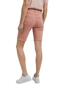 Personne portant un short en jean rose jusqu'aux genoux et des baskets blanches, debout de dos au spectateur.