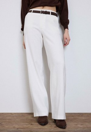 Kobieta ubrana w białe spodnie z wysokim stanem i szerokimi nogawkami, brązowy pasek, brązowy sweter i brązowe buty w szpic stoi na drewnianej podłodze.