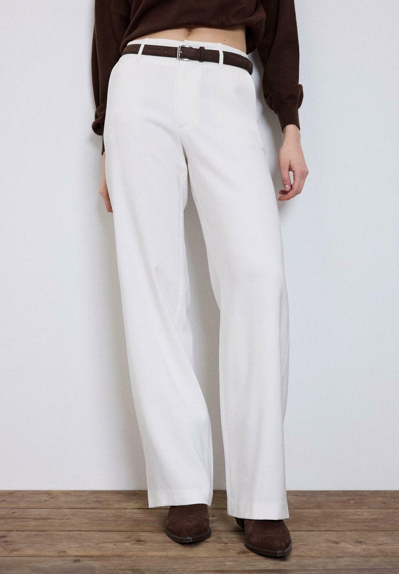 Femme portant un pantalon blanc taille haute à jambes larges, ceinture marron, pull marron et chaussures marron à bouts pointus, debout sur un sol en bois.
