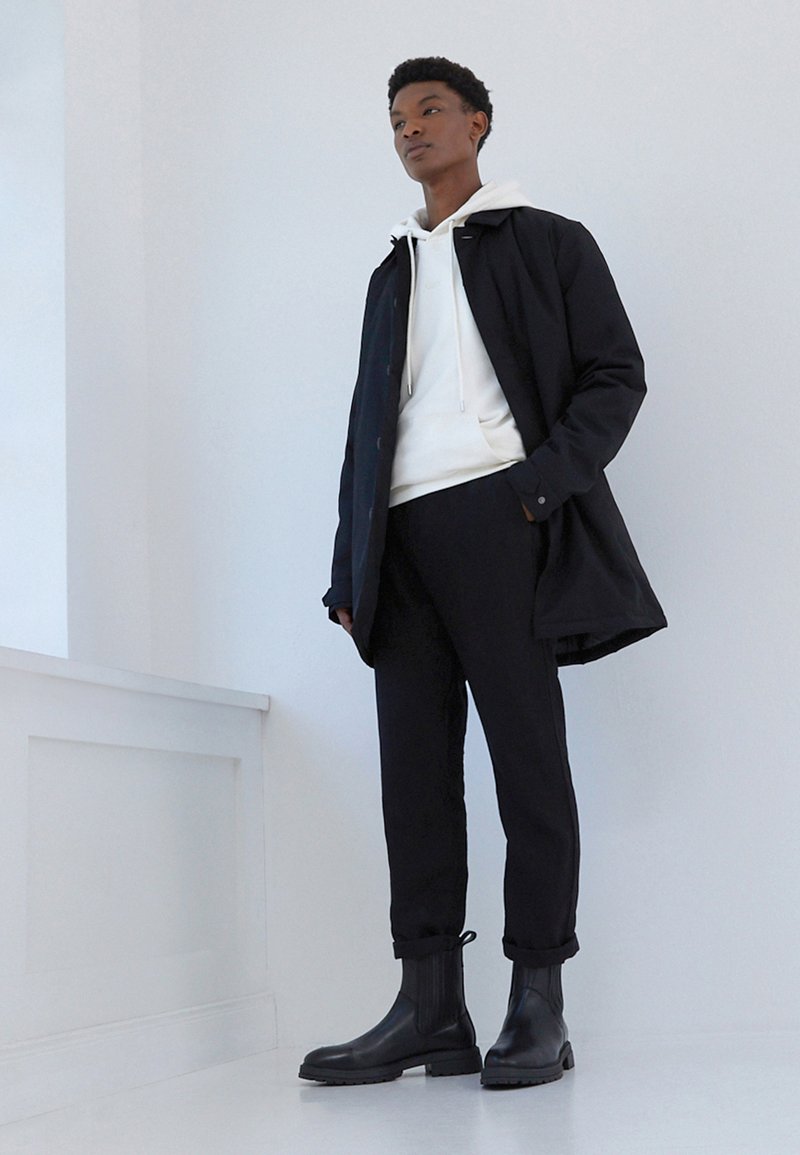 Cappotto nero sopra una felpa bianca, abbinato a pantaloni neri e stivaletti neri lucidi. Texture morbide e design minimalista.