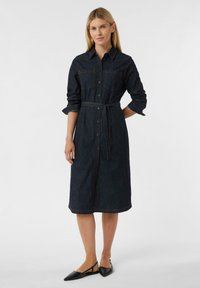 Sukienka koszulowa z denimu w ciemnoniebieskim kolorze, z guzikami, długimi rękawami i paskiem w talii do związania. Posiada dwie kieszenie na piersi oraz kontrastowe przeszycia.