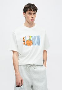 Λευκό βαμβακερό μπλουζάκι με το γράφημα "AIR JORDAN" που συνδυάζει στοιχεία μπάσκετ και καρτούν. Κοντά μανίκια, άνετη εφαρμογή, συνοδευόμενο από γκρι σορτς.