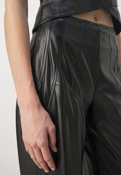 Fekete bőr nadrág fényes felülettel, bonyolult varrásokkal és magas derékkal. A csípőn pihenő kéz kiemeli a textúrát.