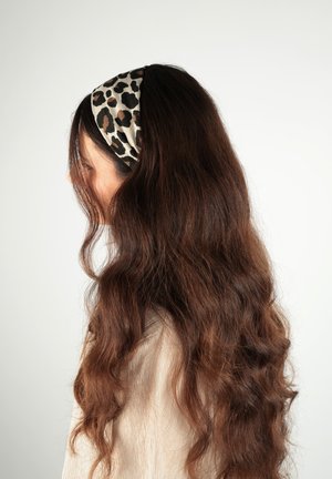 Donna con capelli castani lunghi e ondulati che indossa una fascia per capelli a stampa leopardata, rivolta a sinistra su uno sfondo chiaro e semplice.