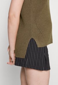 Olivgrön stickad topp med rundad nederkant och sidofickor, ihop med svarta randiga shorts. Texturerad tyg med ribbade detaljer.