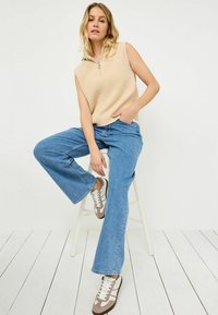 Beige ärmlös tröja med dragkedja, ljusblå utsvängda jeans och bruna sneakers med vita snören. Modell sitter på en vit pall.