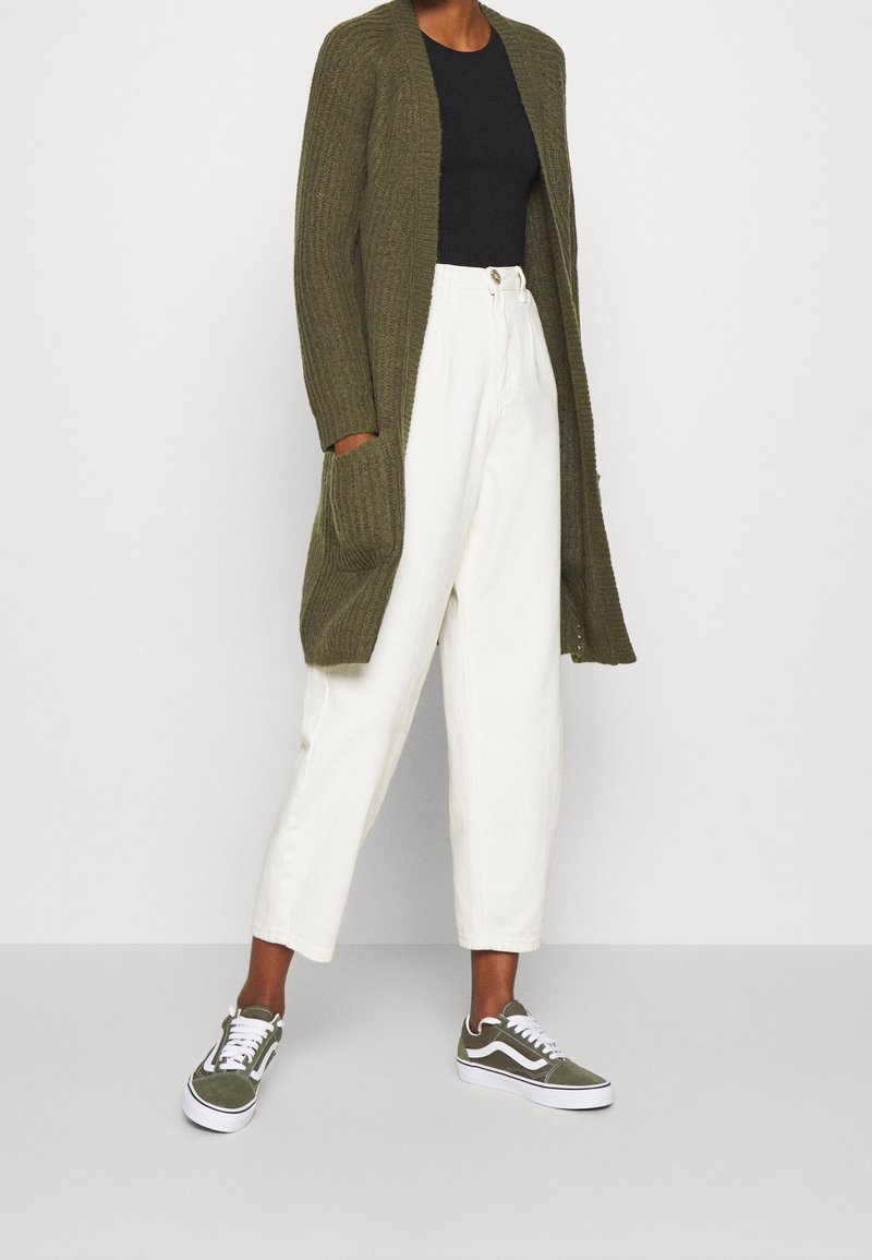 Personne portant un long cardigan vert olive, un top noir, un pantalon blanc taille haute et des baskets vert olive, debout sur un fond blanc.