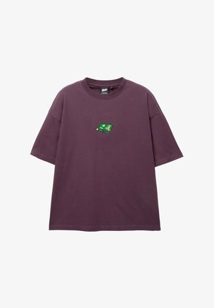 Fioletowa bawełniana koszulka z luźnym krojem, z małą zielono-żółtą haftowaną naszywką na klatce piersiowej. Krótkie rękawy i okrągły dekolt.