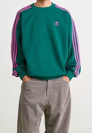 Mężczyzna w zielonej bluzie Adidas z różowymi paskami na rękawach i szarych spodniach sztruksowych, z rękami w kieszeniach.