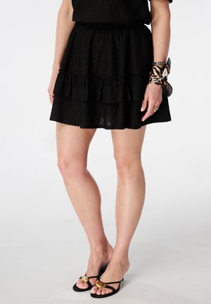 Femme portant une mini-jupe noire texturée à volants, des sandales noires sans lacets avec des accents dorés, et une écharpe imprimé léopard nouée autour de son poignet.
