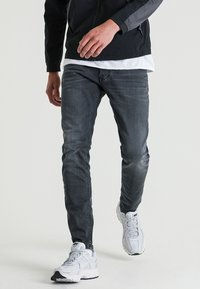 Jean en denim gris foncé avec une coupe slim, présentant un léger effet délavé. Associé à des baskets blanches de sport et à une veste noire. Design simple.