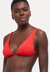 Crveni bikini top s trokutastim dizajnom, glatkom teksturom i tankim trakama. Ključne značajke uključuju dubok V-izrez i minimalne detalje šivanja.