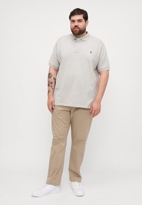 Polo cinza claro com mangas curtas e colarinho, combinado com calças chino bege e sapatilhas brancas, apresentando um pequeno logótipo verde no peito.