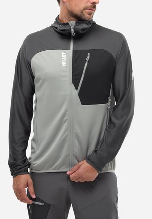 Homme portant une veste zippée bicolore gris et noir avec des poches zippées sur la poitrine et sur les côtés, regardant sur le côté en ajustant l'ourlet avec la main.