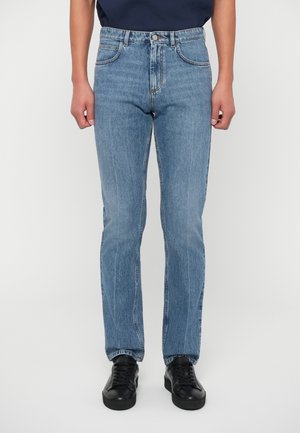 Person, der gerade geschnittene blaue Jeans und schwarze Schnürschuhe trägt, steht vor einem schlichten weißen Hintergrund.