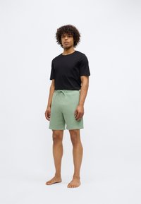 Tricou negru cu mânecă scurtă purtat împreună cu pantaloni scurți texturați, de culoare verde deschis, cu talie elastică, prezentat pe un model desculț.