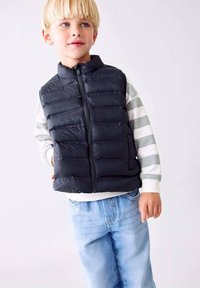 Čierny puff vest s horizontálne prešívanými panelmi, s predným zipsom, nosený cez šedobielo pruhovaný dlhý rukáv tričko, skombinovaný so svetlomodrými džínsami.