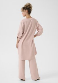 Tunică roz deschis cu mâneci lungi și hem rotund, asociată cu pantaloni largi asortați. Textură moale, design simplu și accente cu nasturi la mâneci.