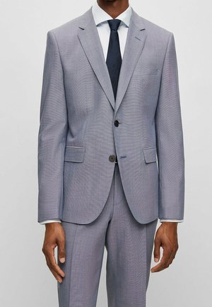 Man in lichtgrijs pak met tweeknopjes jasje, wit overhemd en donkerblauwe stropdas, getoond van nek tot halverwege de dij tegen een effen achtergrond.