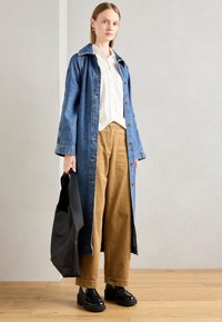Vrouw die binnen staat, gekleed in een lange blauwe denimjas, een witte button-up shirt, een beige corduroybroek, zwarte loafers, en een grote zwarte tote bag vasthoudt.