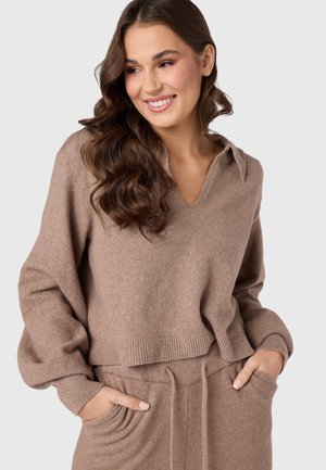 Beige gebreide trui met V-hals, lange mouwen en geribbelde zoom, gecombineerd met bijpassende broek met trekkoord in de taille, die een zachte textuur laat zien.