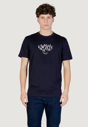 Giovane uomo con capelli ricci che indossa una T-shirt Armani Exchange blu navy e jeans scuri, in piedi contro uno sfondo chiaro semplice.