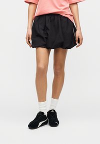 Personne portant un t-shirt rose, un short bouffant noir, des chaussettes blanches et des baskets Puma noires, debout devant un fond uni.