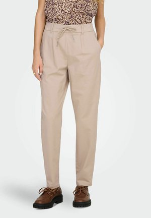 Kobieta ubrana w beżowe spodnie o zwężanych nogawkach z sznurkiem w pasie, brązową, wzorzystą bluzkę oraz brązowe wiązane buty, stojąca z jedną ręką w kieszeni.