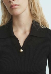 Top noir à côtes avec un col classique, présentant un détail de bouton doré au niveau du col. Texture douce et design ajusté.