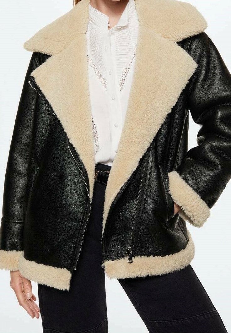 Veste en cuir noire avec doublure en shearling beige au col, aux poignets et à l'ourlet portée sur une chemise blanche texturée à boutons et un pantalon noir.