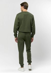 Olīvu zaļš sporta tērps, kas sastāv no pulovera ar apaļu kaklu un sašaurinātām biksēm. Izgatavots no mīksta, strukturēta auduma ar ribu aprocēm un jostasvietu.