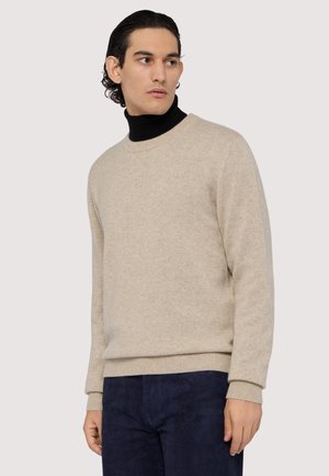 Beige strikbluse med rund hals og ribbede manchetter, båret over en sort turtleneck, parret med mørk navy bukser. Blød tekstur.