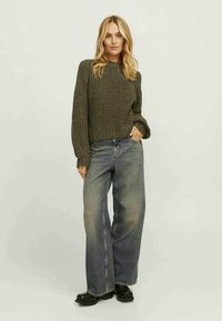 Suéter verde-azeitona, de ponto grosso, com mangas longas, combinado com jeans azuis de perna larga e alta cintura, e calçado preto.