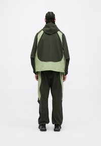 Groen en lichtgroen sportoutfit in twee delen, met een hoodie-jack met een grote ruginzet en ontspannen pasvorm broek. Zwarte schoenen.