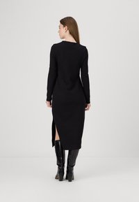 Vestido preto de manga longa com uma silhueta justa, fendas laterais e uma textura suave. Combinado com botas altas até ao joelho brilhantes.