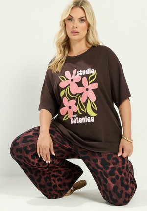 Kvinde med langt blond hår iført en brun T-shirt med pink blomster og tekst, kombineret med vidde bukser med leopardprint, knælende på det ene knæ.