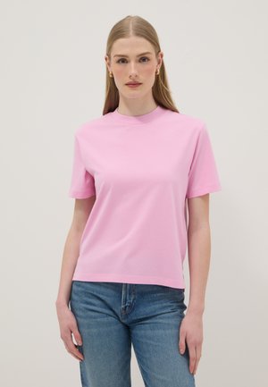Femme aux longs cheveux blonds, portant des boucles d'oreilles créoles dorées, un t-shirt rose à manches courtes et un jean bleu, debout devant un fond clair uni.