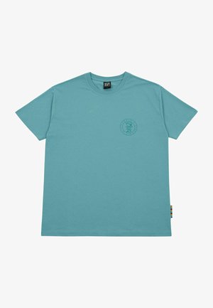 Gaiši zils kokvilnas T-krekls ar īsām piedurknēm, apaļu kaklu un apļa grafisku logo uz krūtīm. Sānu šuvē ir multicolora etiķete.