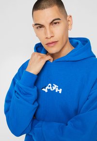 Modra bombažna kapuca z belim grafičnim napisom "Mladost" na prsih. Kapuca z vrvico in rebrastimi manšeti.