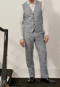 Gilet a scacchi grigi e pantaloni abbinati realizzati in tessuto testurizzato, abbinati a una camicia bianca, scarpe nere e dettagli dei bottoni in evidenza.