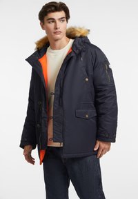 Chaqueta de abrigo navy con capucha forrada de pelo, forro naranja, detalles de botones y bolsillos laterales. El modelo lleva una camisa clara y jeans oscuros.