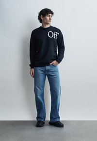 Ung mand med krøllet hår iført en sort sweater med teksten "Off", blå jeans og sorte sko, stående mod en ensfarvet lys grå baggrund.