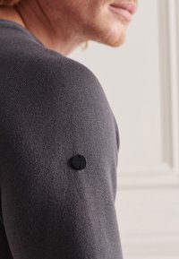 Pull gris en tricot avec une finition texturée, avec une étiquette noire de marque sur l'épaule. Vue en gros plan montrant la forme et les détails du tissu.