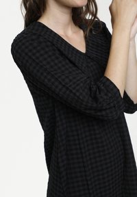 Vestido de tela a cuadros en negro y gris oscuro, con un cuello redondeado, mangas de tres cuartos y detalles fruncidos en los puños de las mangas.