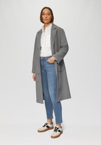 Grijze wollen jas, witte shirt, blauwe jeans en beige en zwarte sneakers. De jas heeft twee voorzakken en een open ontwerp.